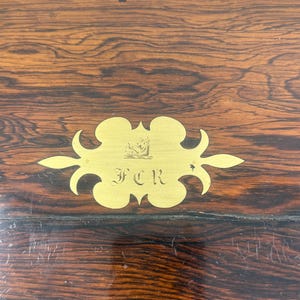 May include: Close-up of a wooden surface with a rich, dark grain pattern. A gold-colored decorative plaque with the initials "FCR" is affixed to the wood. The plaque has an ornate, stylized design.