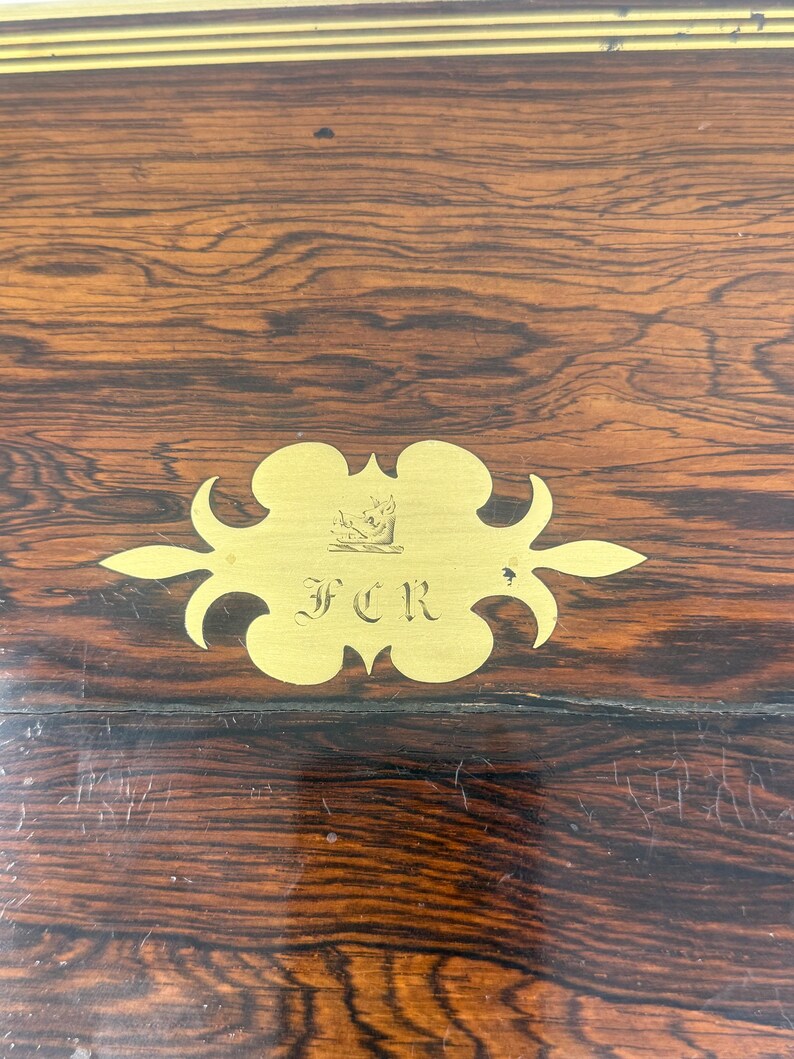 May include: Close-up of a wooden surface with a rich, dark grain pattern. A gold-colored decorative plaque with the initials "FCR" is affixed to the wood. The plaque has an ornate, stylized design.