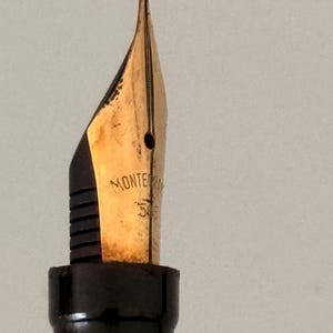 May include: A gold-colored fountain pen nib with a black feed, featuring the engraved text "MONTE" and "585" on the metal surface. The nib has a central breather hole and a vertical slit extending to the tip, attached to a black plastic grip section.