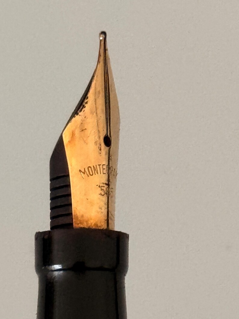 May include: A gold-colored fountain pen nib with a black feed, featuring the engraved text "MONTE" and "585" on the metal surface. The nib has a central breather hole and a vertical slit extending to the tip, attached to a black plastic grip section.