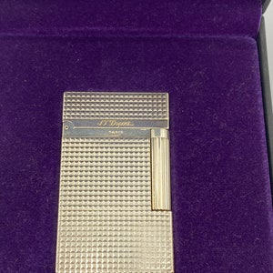 Pode incluir: Isqueiro prateado com um padrão de diamante e o texto "S.T. Dupont Paris" na frente.