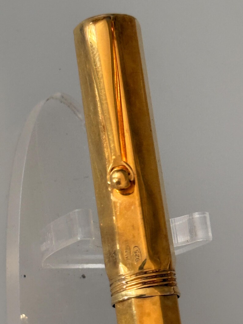 May include: A gold-colored fountain pen cap with a smooth, reflective surface and a rounded clip attachment. The metal surface features a small, stamped hallmark reading 925, indicating sterling silver composition beneath the gold-toned finish.