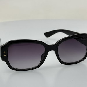 Puede incluir: Gafas de sol negras con lentes gris oscuro y un acento dorado en el lateral. Las gafas tienen una forma rectangular y un logotipo de Dior en la lente derecha.