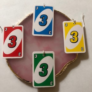 Könnte beinhalten: Vier Uno-Kartenohrringe mit der Zahl 3 in Rot, Blau, Gelb und Grün. Jede Karte hat die Zahl 3 in einem weißen Oval mit weißem Rand.