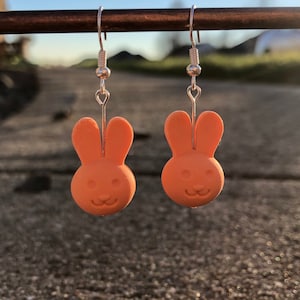 May include: A pair of orange bunny-shaped earrings. The earrings are made of a smooth material and feature a simple design with small eyes and a smiling mouth. The earrings hang from silver-colored hooks.
