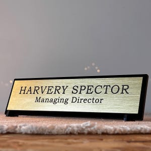 May include: Gold desk nameplate with black lettering that reads "Harvey Spector Managing Director".