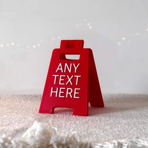Può includere: Un cartello rosso vivo a forma di A con le parole "ANY TEXT HERE" in bianco. Il cartello è realizzato in un materiale solido e poggia su una superficie strutturata bianco sporco. Il cartello è progettato per testo personalizzato.