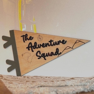 May include: Wooden pennant flag with the text "The Adventure Squad" in black lettering. The flag is decorated with a compass, mountains, and a dotted line.