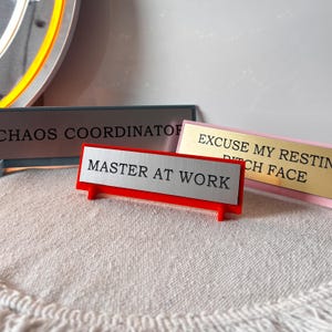 May include: A selection of desk nameplates with amusing inscriptions. The front nameplate says "MASTER AT WORK" on a silver plate with a red base. Other plates read "CHAOS COORDINATOR" and "EXCUSE MY RESTING BITCH FACE".
