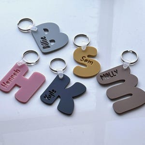 Può includere: Una collezione di portachiavi personalizzati in varie forme e colori. Ogni portachiavi presenta una lettera dell'alfabeto, con nomi come "Billie", "Hannah", "Kyle", "Sam" e "Molly" incisi. I portachiavi sono attaccati a portachiavi argentati.