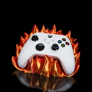 Può includere: Un controller Xbox bianco con pulsanti neri e pulsanti azione colorati poggia su un supporto a forma di fiamma arancione e oro stampato in 3D. Il controller ha il logo Xbox e un D-pad.