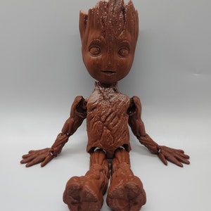 May include: A brown, articulated, 3D printed figurine of a character from the Guardians of the Galaxy movie franchise. The character is sitting with its legs crossed and its arms outstretched. The figurine is detailed with a textured surface and a realistic facial expression.
