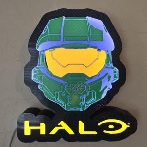 May include: A wall-mounted LED sign featuring the Master Chief helmet from the Halo video game series. The helmet is illuminated in green and blue with a bright yellow visor. Below the helmet, the word HALO is displayed in yellow text.