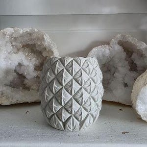 May include: A light gray cylindrical planter featuring a repeating geometric diamond pattern across its exterior surface. The planter has a textured, matte finish and is positioned in front of two large, white, crystalline rock formations.