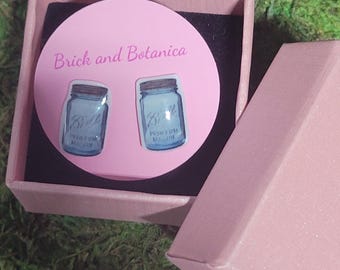 May include: A pink box with a white circle inside. The circle has the text "Brick and Botanica" at the top and two blue mason jar earrings with the text "Ball" on them.