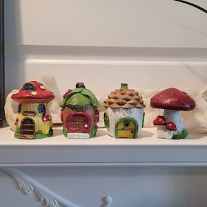 May include: Four miniature fairy houses made of painted resin. The houses are shaped like mushrooms, a cottage with a green roof, a cottage with a brown roof, and a mushroom with a red cap. The houses are all decorated with details like windows, doors, and chimneys.