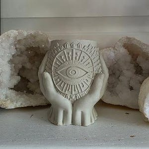 May include: A light gray concrete planter shaped like two cupped hands holding a circular vessel. The front of the vessel features a detailed embossed design of an eye surrounded by radiating sunburst lines and geometric patterns.