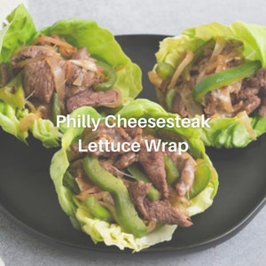 Puede incluir: Tres wraps de lechuga rellenos de filete de ternera cortado fino, pimientos verdes y cebollas. Los wraps est&aacute;n en un plato negro y el texto "Philly Cheesesteak Lettuce Wrap" est&aacute; escrito debajo de los wraps.