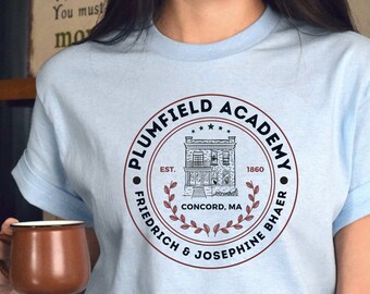 Op de afbeelding: Een lichtblauw t-shirt met een cirkelvormig grafisch ontwerp met een gebouw, tekst en een lauwerkrans. De tekst luidt "Plumfield Academy", "Est. 1860", "Concord, MA", "Friedrich & Josephine Bhaer".