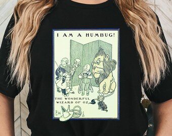 May include: Black t-shirt featuring a vintage illustration from "The Wonderful Wizard of Oz." The artwork includes characters like the Lion, Scarecrow, Tin Man, and Dorothy, with the text "I AM A HUMBUG!" above the image.