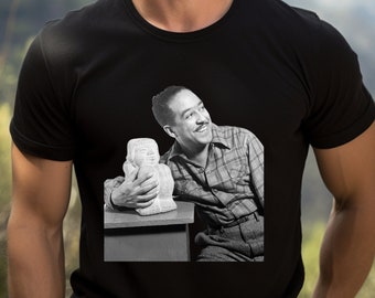 Puede incluir: Camiseta negra con una imagen en blanco y negro de un hombre sonriendo, sosteniendo una escultura. El hombre lleva una camisa a cuadros y pantalones. La escultura es de color claro y parece ser un busto.