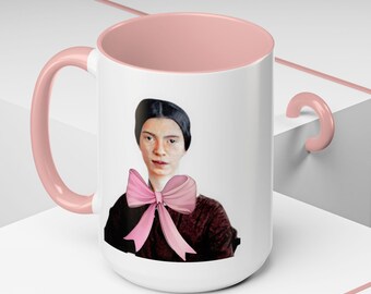 Può includere: Una tazza in ceramica bianca con interno e manico rosa. La tazza presenta un ritratto di una donna con un fiocco rosa intorno al collo.