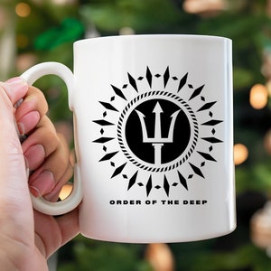 May include: White ceramic mug with a black and white graphic of a trident inside a circle with a rope design. The circle is surrounded by a ring of black diamond shapes. The text "ORDER OF THE DEEP" is printed below the graphic.