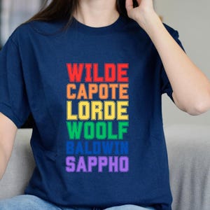 May include: A navy blue t-shirt featuring the names Wilde, Capote, Lorde, Woolf, Baldwin, and Sappho in a rainbow gradient. The text is arranged vertically, each name in a different colour.