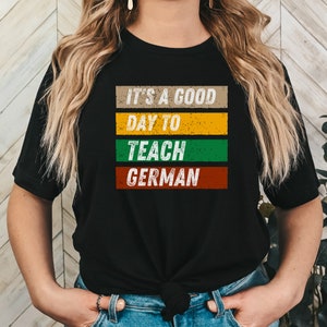 Op de afbeelding: Een zwart T-shirt met een vintage-stijl grafische afbeelding met de tekst "It's a good day to teach German". De afbeelding is in een meerkleurig gestreept ontwerp met een verweerde look.
