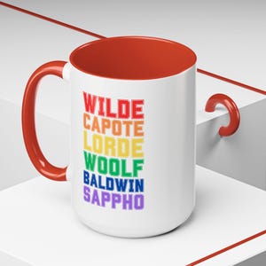 Könnte beinhalten: Eine weiße Keramiktasse mit rotem Henkel und rotem Innenraum. Die Tasse hat ein Regenbogen-Design mit den Namen "WILDE", "CAPOTE", "LORDE", "WOOLF", "BALDWIN" und "SAPPHO" in Schwarz gedruckt.