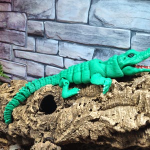 May include: A green plastic toy alligator with a segmented body and an open mouth. The alligator is posed on a piece of bark.