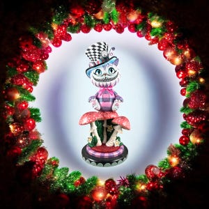 Op de afbeelding: Een grillig beeldje van de Cheshire Cat uit Alice in Wonderland, met een geruite hoge hoed en zittend op rood gestippelde paddenstoelen. De kat heeft een brede grijns en wordt omringd door een kerstkrans met rode ballen en groen gebladerte.