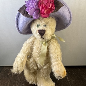 May include: A white teddy bear wearing a lavender hat with pink and purple flowers. The bear has green eyes and a brown nose.