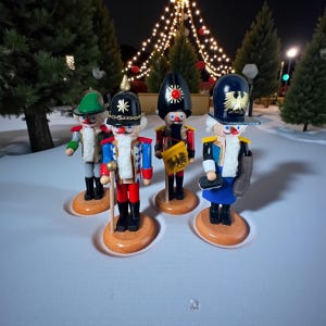May include: Four wooden nutcrackers in soldier uniforms stand on a snowy surface. Each figure has a unique hat and uniform design, with details like gold accents, a star, and a flag. The background features Christmas lights and trees.