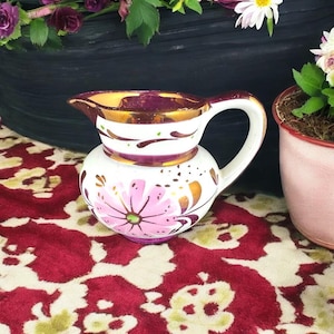 May include: A small white ceramic jug with a pink and gold floral design. The jug has a rounded body and a handle. The jug is sitting on a red and white patterned surface.