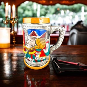 Peut inclure: Une chope à bière en céramique avec une illustration colorée d'un roi de carte à jouer. La chope a une poignée et un bord jaune.