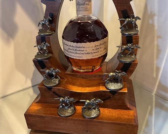 Puede incluir: Una botella de whisky bourbon Blanton's Single Barrel exhibida en un soporte de madera en forma de herradura. El soporte presenta figuras en miniatura de caballos y jinetes y está hecho de madera oscura. La botella tiene un tapón de metal con un caballo y un jinete.