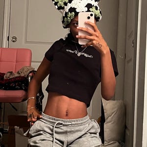 Op de afbeelding: Een gehaakte hoed met een koeienpatroon in zwart, wit en groen. De hoed wordt gedragen met een zwarte crop top en een grijze joggingbroek. Een roze stoel en een waterfles zijn ook zichtbaar.