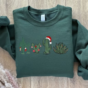 May include: A dark green sweatshirt with a Christmas cactus design. The cactus is wearing a Santa hat and has Christmas lights and ornaments.