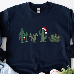 May include: A navy blue sweatshirt with a Christmas cactus design. The cactus is wearing a Santa hat and has Christmas lights and ornaments.