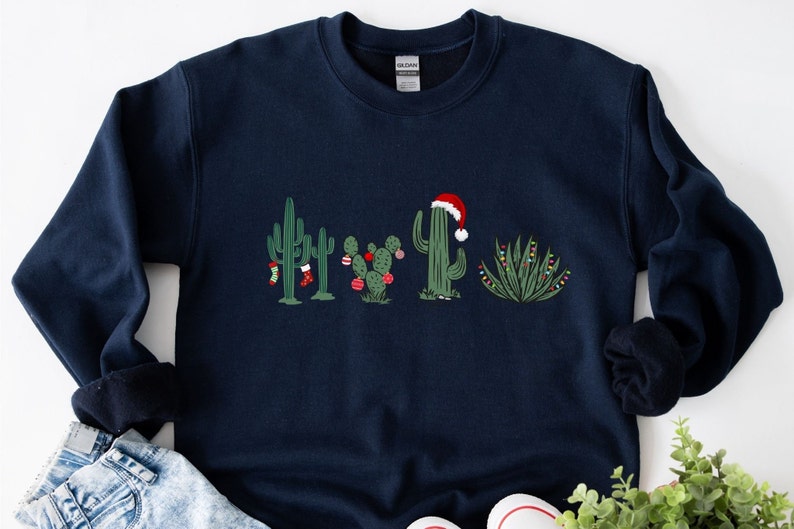 May include: A navy blue sweatshirt with a Christmas cactus design. The cactus is wearing a Santa hat and has Christmas lights and ornaments.