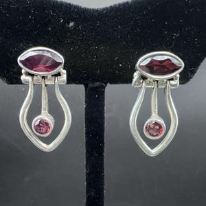 May include: A pair of silver earrings featuring a marquise-cut deep red gemstone at the top and a smaller round-cut red gemstone suspended within a teardrop-shaped silver frame. The metal has a polished finish and the design is symmetrical.