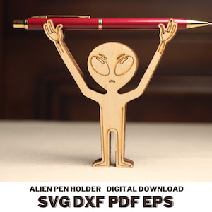 May include: A wooden alien pen holder with outstretched arms holding a red pen. The alien figure has large oval eyes and a small mouth. The text "ALIEN PEN HOLDER DIGITAL DOWNLOAD SVG DXF PDF EPS" is at the bottom.