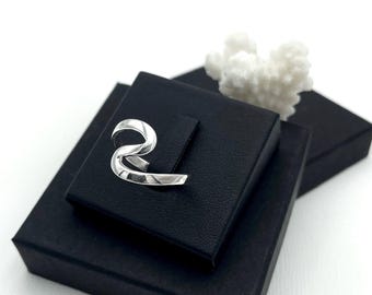 May include: A silver-colored ear cuff with a curved, abstract design. The ear cuff is displayed on a black box, with a white coral-like object in the background. The image is set against a white backdrop.