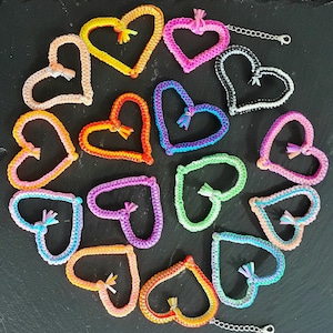 May include: A collection of 14 colorful crocheted heart-shaped keychains with tassels. The keychains are arranged in a circle on a black background.