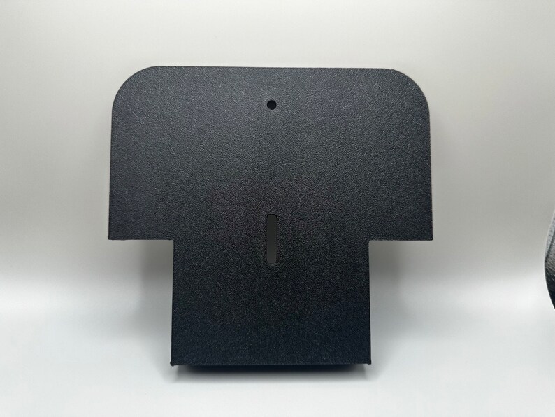 May include: Black plastic wall mount with a rounded top and a rectangular opening in the center. The mount has a textured surface.