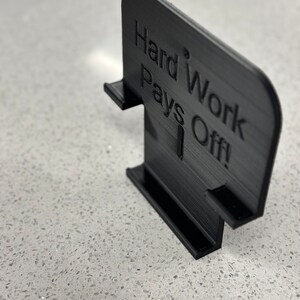 May include: Black 3D printed sign with the text "Hard Work Pays Off!" The sign is designed to stand upright on a flat surface.