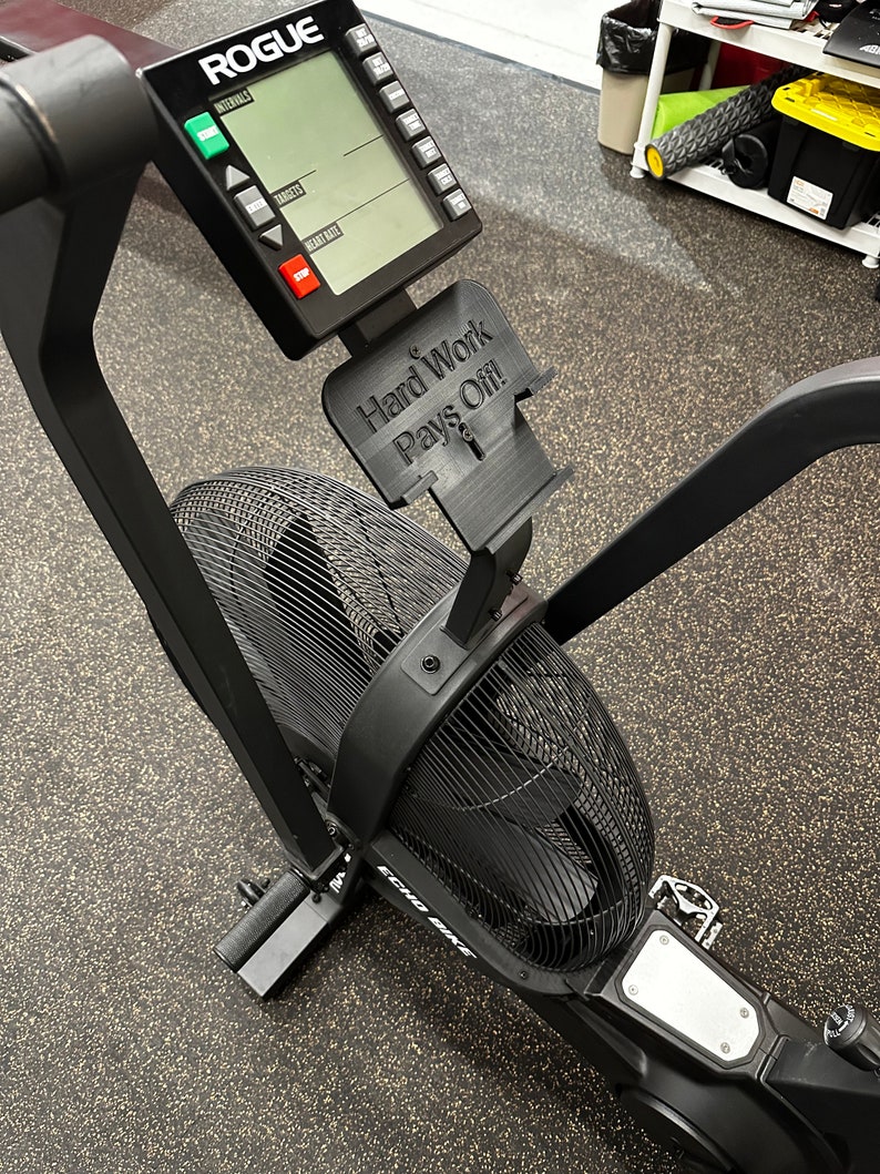 May include: Black air bike with a digital display and a motivational sign that reads "Hard Work Pays Off!" The bike is on a black rubber mat.