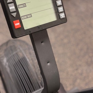 May include: Close-up of a black rowing machine console with a digital display showing the words "Targets", "Heart Rate", "Custom", "Target Time", "Target Dist", "Target Cals", and "Target HR". The console has buttons for "Start", "Enter", "Stop", and other functions.