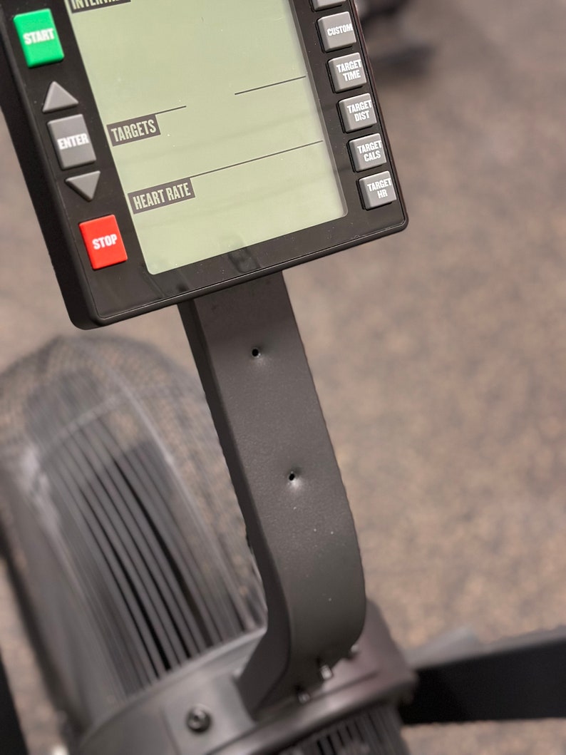 May include: Close-up of a black rowing machine console with a digital display showing the words "Targets", "Heart Rate", "Custom", "Target Time", "Target Dist", "Target Cals", and "Target HR". The console has buttons for "Start", "Enter", "Stop", and other functions.